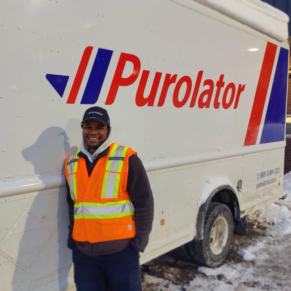 Tyrel Rojan‑Jackson smiling beside a Purolator courier truck wearing a high‑visibility vest and Purolator branded hat.